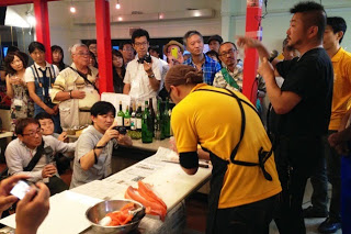 黄色いシャツを着た料理人がサーモンのような魚をさばき、その周りを多くの参加者が囲んで見守っている、魚のさばき方の実演を見学している様子の写真