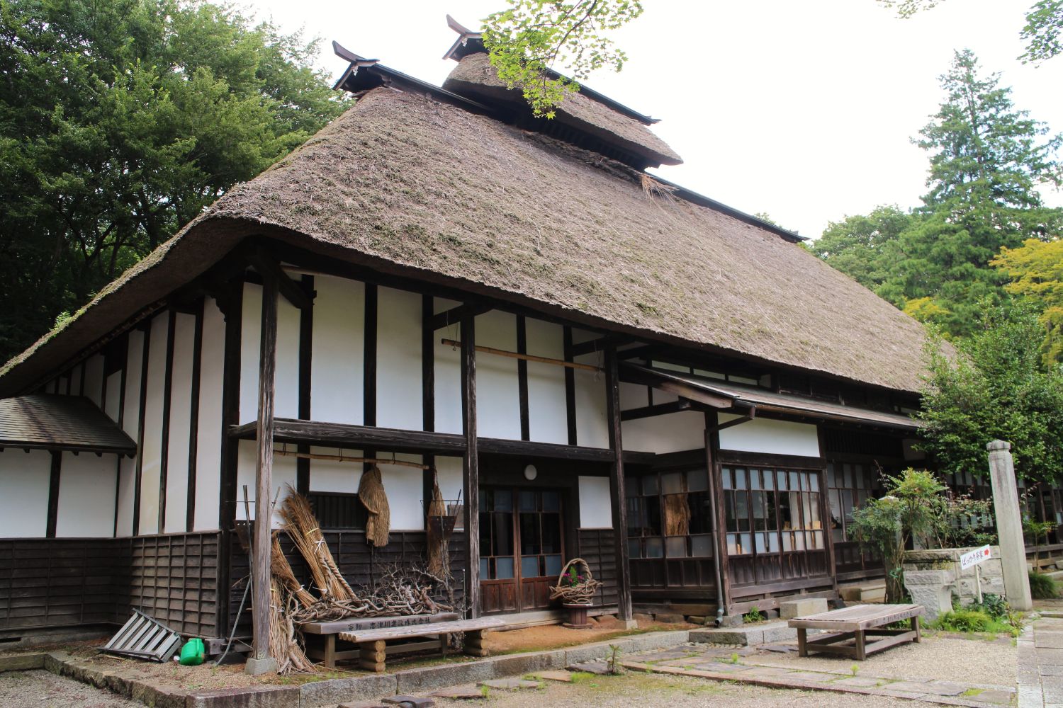 茅葺き屋根と白壁の伝統的な日本の古民家が、木々に囲まれた静かな場所に建っている様子の写真