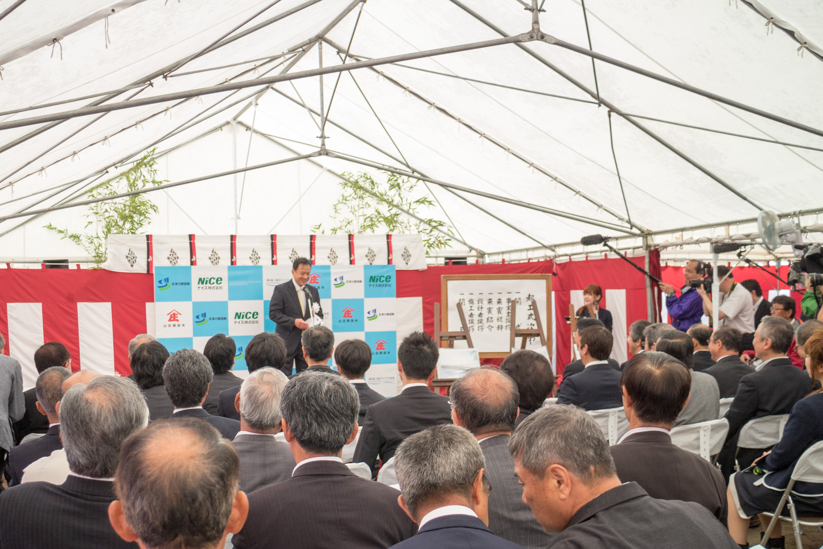 白いテント内で多くのスーツ姿の男性たちが椅子に座って前方の壇上で話す男性の話を真剣に聞いている起工式の式典の様子を写した写真