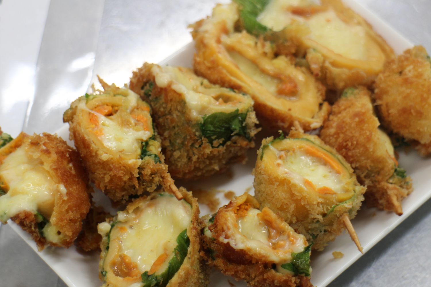 ホヤにチーズをたっぷり詰めて大葉を巻き、爪楊枝をさし衣をつけて揚げた料理が、カットされた状態で皿に盛られている「ホヤチーズフライ」の写真