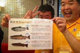 黄色いシャツを着た男性が笑顔で「利きサケ」の説明が書かれた用紙を楽しそうに手に持っている写真