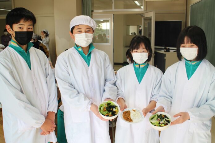 4人の中学生の生徒たちが緑のジャージの上に白衣とマスクを着け、右側の3人の生徒が手にそれぞれ料理の載った皿を持ってカメラに向かって並んで立っている写真