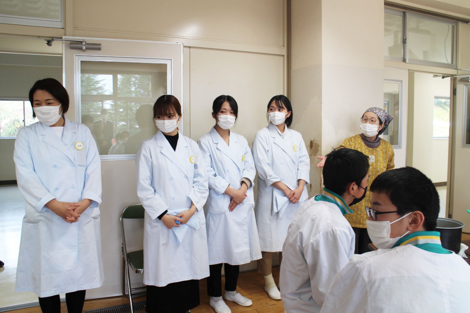 白衣を着用した4人の女性が壁の前に並んで立ち、右端に立っている割烹着を着た三角巾着用の女性が右手を出して生徒たちに向かって紹介をしている様子の写真