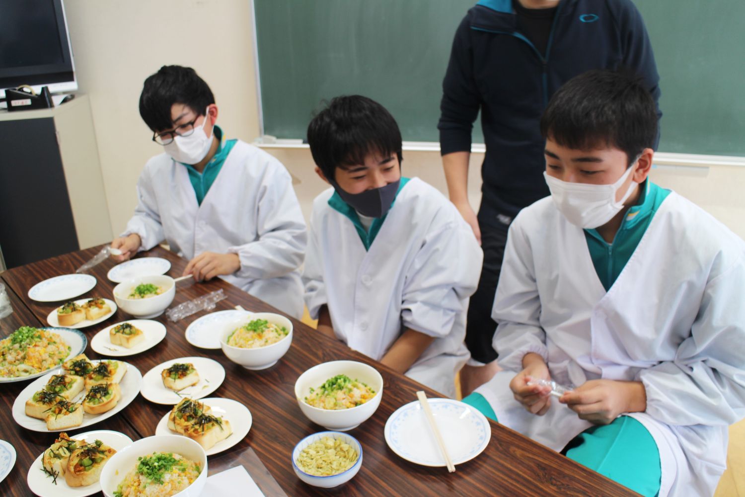 給食用の白衣を着た3人の男子生徒が、机に並べられた沢山のホヤ料理を前にして座っている試食の様子の写真