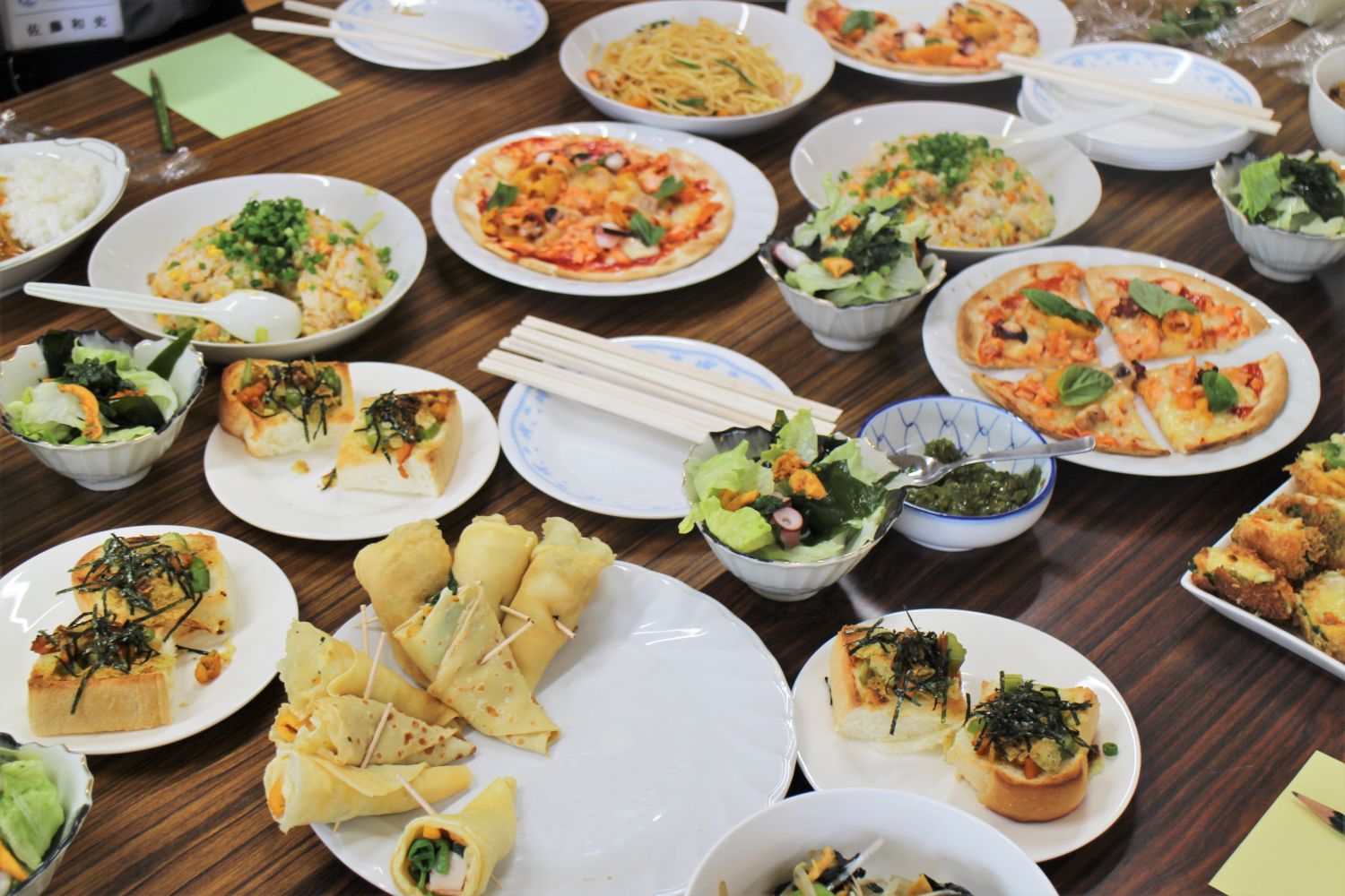 木目の机の上に、ホヤを使用したピザやチャーハン、サラダ、クレープ、パンなど様々な料理が並んでいる写真