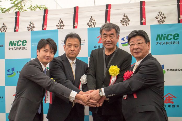 スーツ姿の男性4人が白と水色の配色のバックパネルの前で手を重ねて握手をしながらカメラに向かって並んでポーズを取っている記念撮影の写真