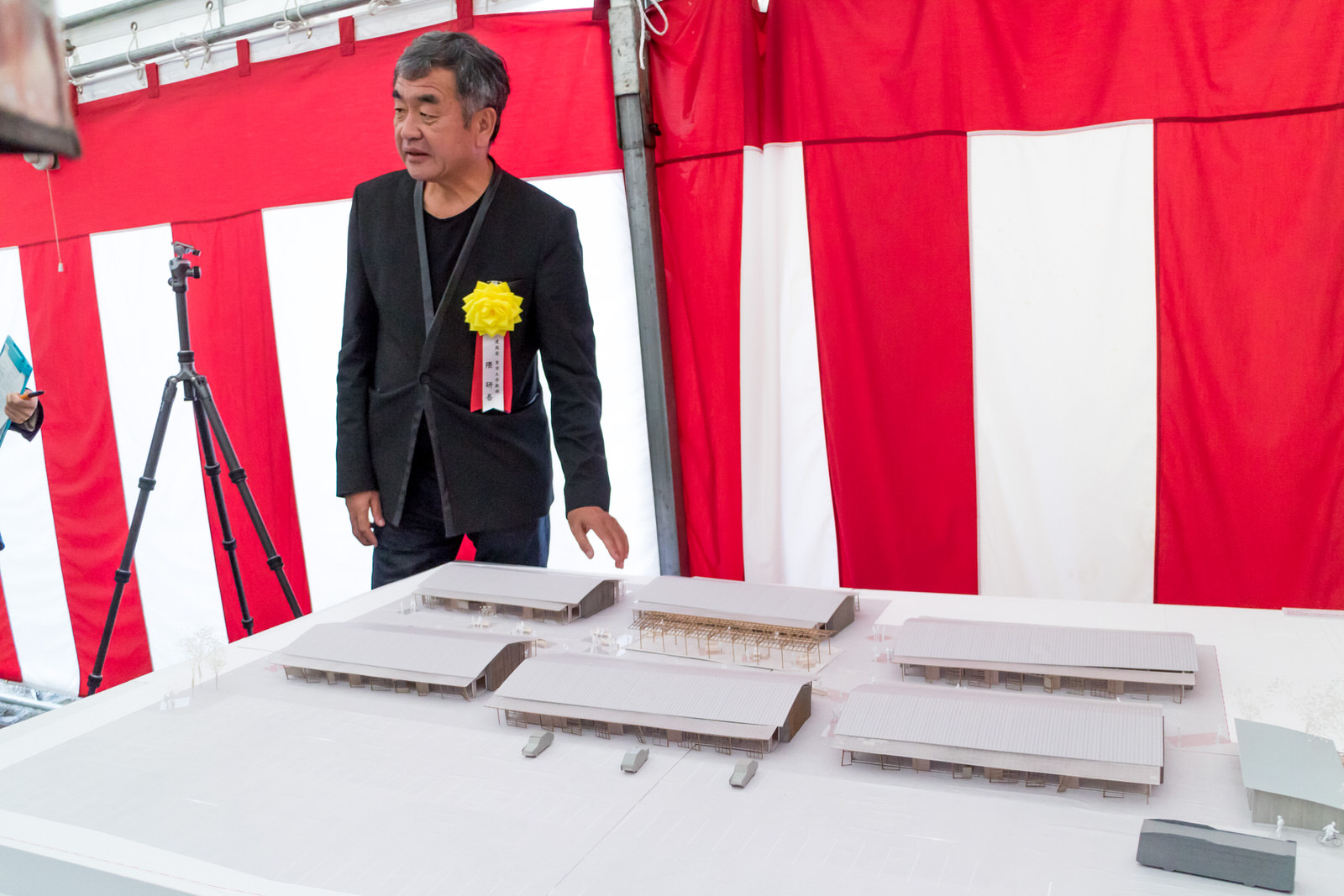 黒のジャケットに大きな黄色いリボンをつけた隈研吾さんが赤白の幕の前で、さんさん商店街の白い模型を並べたテーブルの横に立って説明している写真