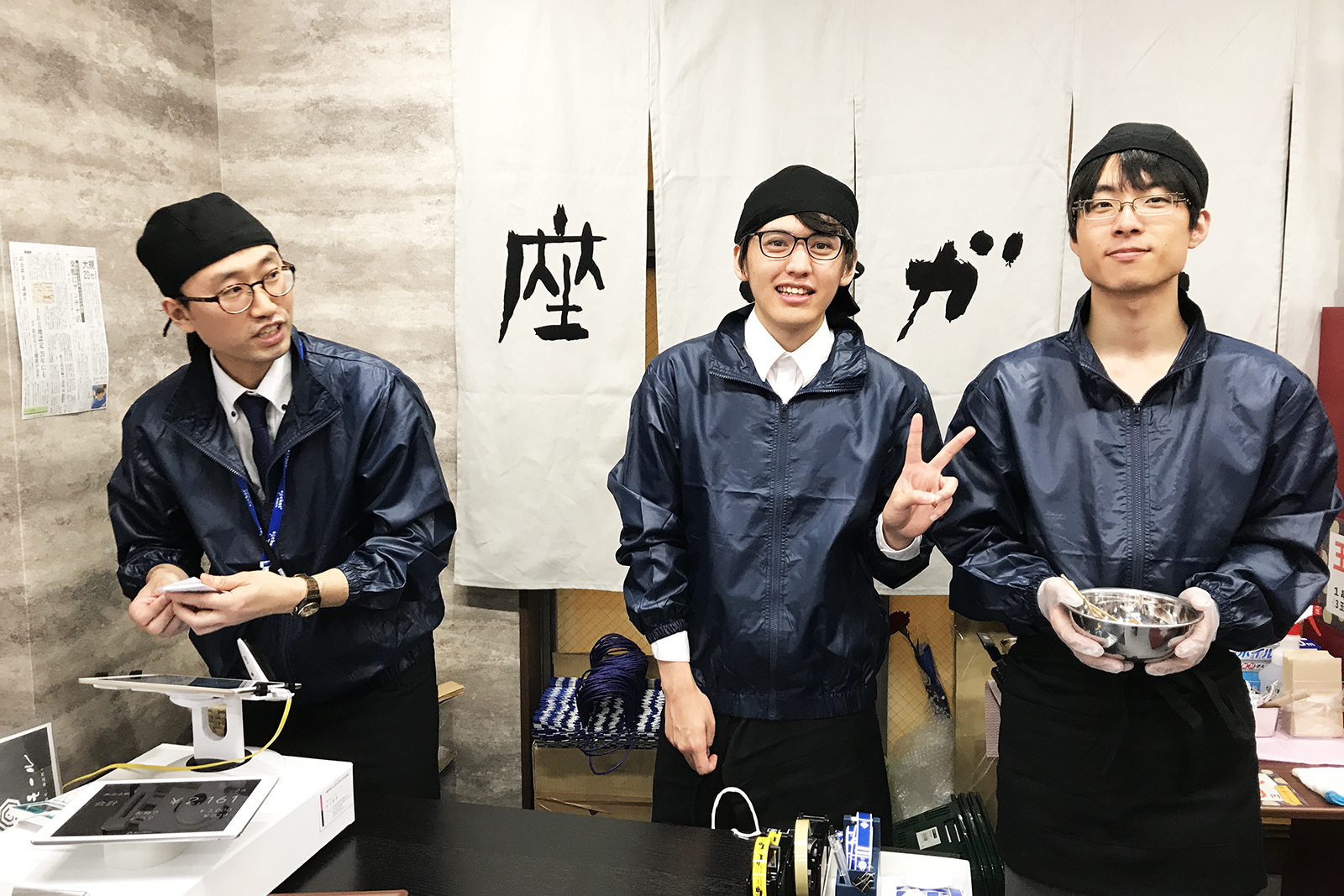 おそろいの紺色の上着と黒い帽子を着用した男性スタッフ3人が笑顔でカウンターの中に立っている写真