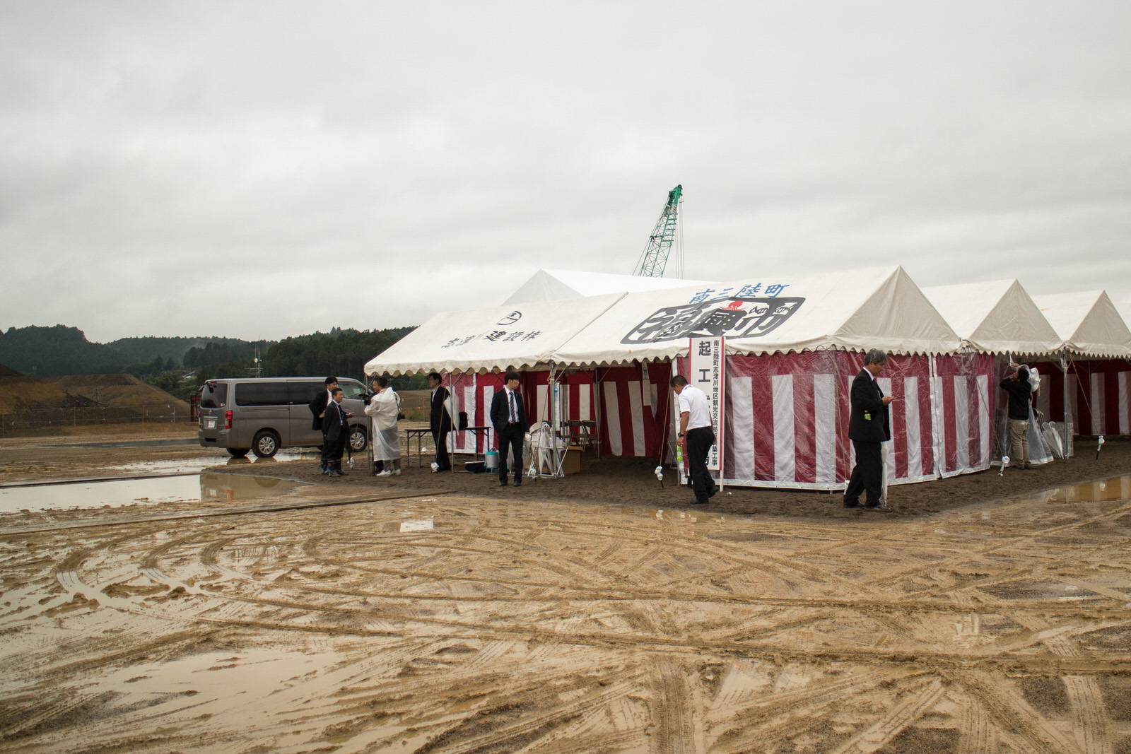 雨でぬかるんだ工事現場に設置された赤白幕のテントの前で、スーツ姿の人々が立ち話や準備をしている起工式会場を写した写真