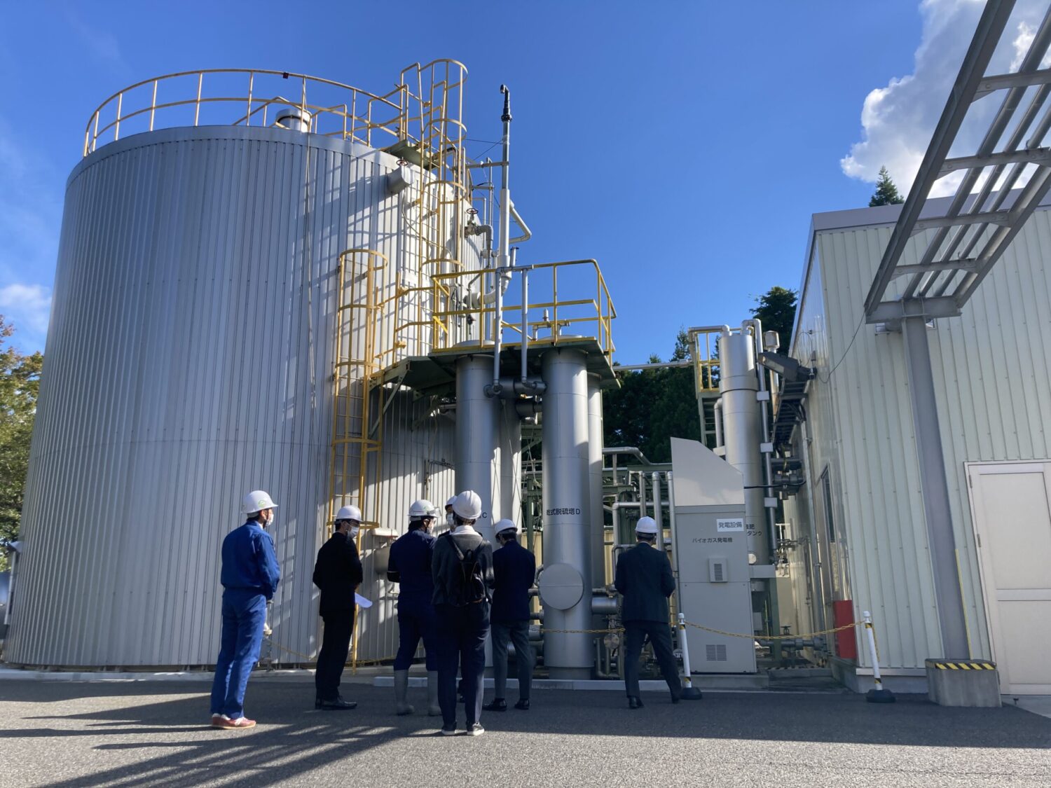 銀色で縦に長い大型の円筒形タンクやパイプ、黄色の手すりと階段が備え付けられたバイオガス施設の前でヘルメットをかぶった町の関係者や見学者たちがその設備を見上げながら説明を受けている、再生可能エネルギーの現場視察の写真