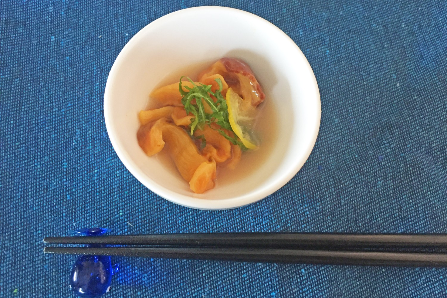 小さな白い椀にスライスされたホヤの身を盛り付け、大葉と薄切りにカットしたレモンを飾り、出汁に浸したホヤの刺身に箸置きと箸が添えられている写