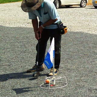 麦わら帽子をかぶった男性がクラブでボールを打ち、旗の立つホールポストにボールが入っている瞬間の写真