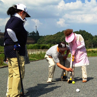 仲間の女性が周囲で見守るなか、男性がピンクの服を着た女性にクラブの使い方を丁寧に教えている写真