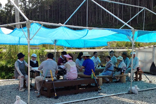 日差しを避けるために青いシートの屋根の下で、十数人の高齢者が長椅子に座って休憩している様子の写真