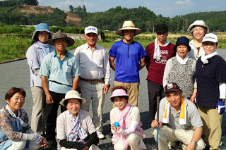 木々が立ち並んだ小高い山の麓の屋外コートに、帽子をかぶった笑顔の参加者たちが2列に並んでいる集合写真