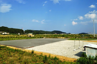 空の下に広がる静かな更地の運動場で、右側には白い砂利が敷かれ、周囲には緑の草木が生い茂っている写真