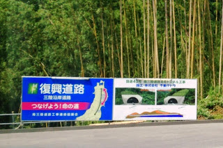 トンネルの完成予想図も描かれている「復興道路 三陸沿岸道路 つなげよう命の道」と書かれた看板が竹林の前に設置された写真