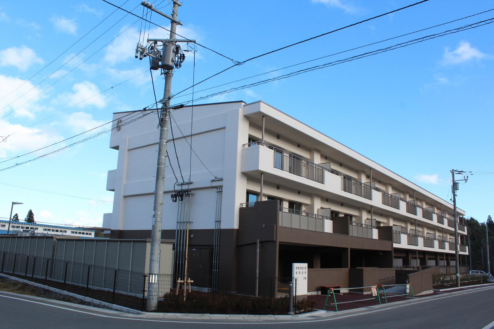 青空の下、「町営志津川西 復興住宅 C棟」と書かれた看板が掲げられた白と茶色の3階建て集合住宅が整備され、舗装された道路やフェンス、植栽なども整った完成間近の様子が写された写真