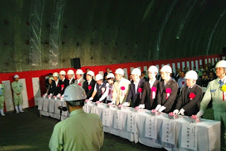 白いヘルメットをかぶり、赤いリボンを胸につけたスーツ姿の関係者たちが白い台に並んで立ち、トンネル貫通式が行われている様子の写真