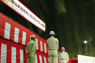 横断幕と紅白幕が飾られたトンネル内で、作業服姿の男性2人と1人が向かい合って並んでいる、開通を祝う式典の様子の写真
