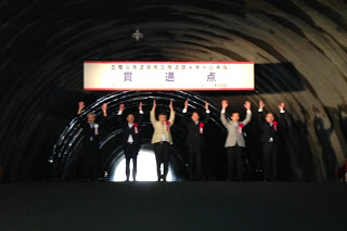 トンネルの貫通点の横断幕の前で、横一列に並んだ6人のスーツ姿の関係者たちが両手を上げて万歳三唱を行っている様子の写真