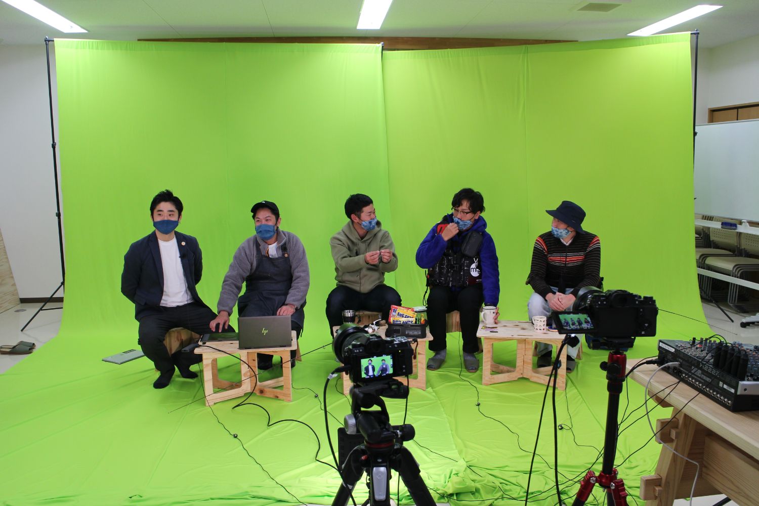 緑のクロマキーの前で5人の男性が並んで座り、マスクを着用して談笑している撮影現場の様子の写真