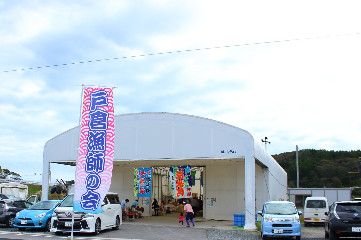 白いテントのような建物の前に複数の車が停まっていて、建物の内部には色とりどりの旗が飾られ、左側には「戸倉漁師の会」と書かれた旗が立っている写真