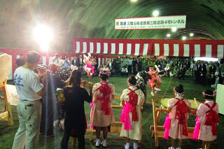 赤白の幕と式典看板が掲げられているトンネル内で、観客が見守る中太鼓演奏と韮の浜獅子舞が披露されている様子を後方から撮影した写真