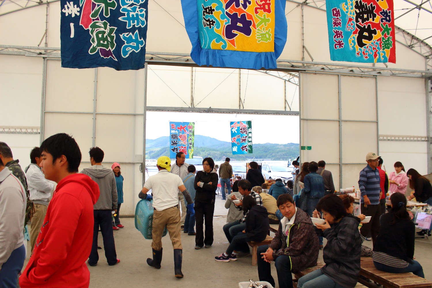 屋根のある広々とした空間で、多くの人々が集まり、座っている人や立っている人が混在しており、上部には色とりどりの旗が吊るされている第32回戸倉漁師の会の様子を写した写真