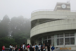 霧に包まれた白い外壁の建物の前に、参加者たちが並び、説明を受けている様子の写真