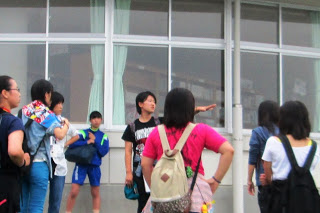 学校の校舎の前で、1人の生徒が手を指しながら他の参加者たちに説明をしている様子の写真