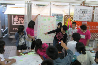 子どもたちのグループが大きな模造紙を広げて発表し、ほかの参加者がその内容を見ているまちづくりワークショップの写真