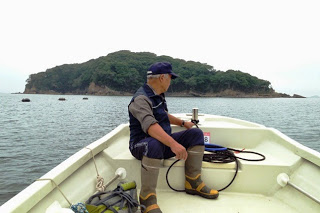 小型船の先頭に腰掛けた紺色のキャップを被った男性が、目前に見える緑に覆われた無人島を眺めている写真