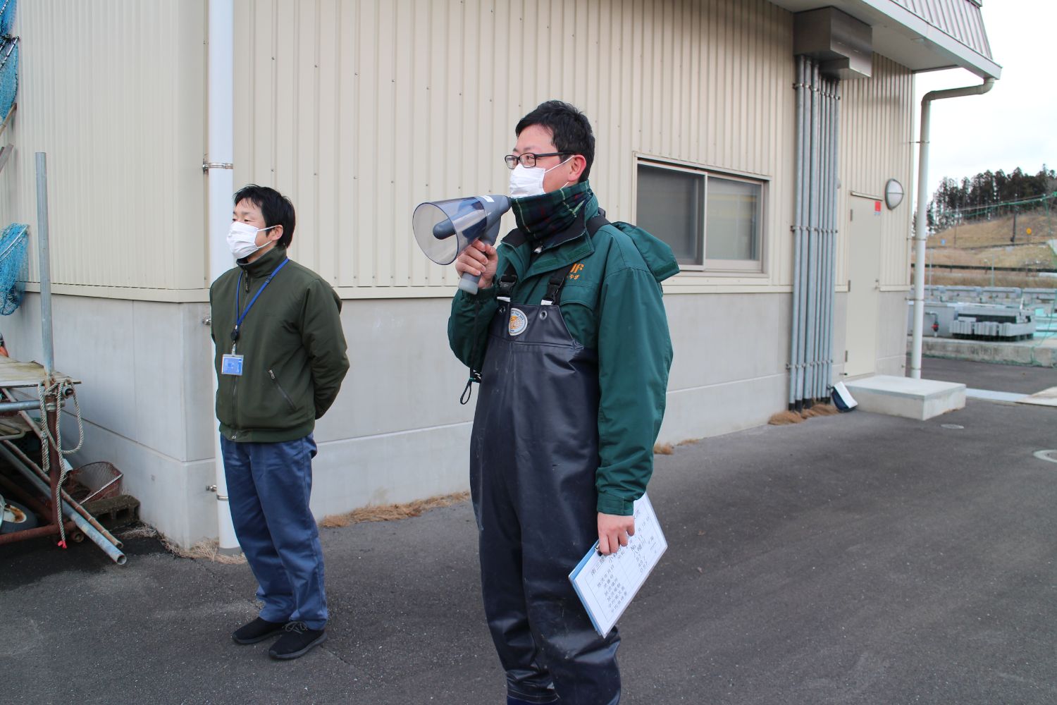 建物の横に一人の男性が立っており、その隣に立った千葉純一さんが拡声器を使って話をしている写真