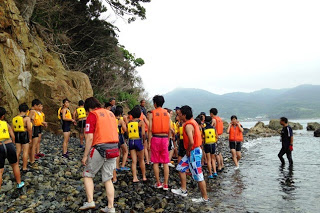 救命胴衣を着た多くの子どもたちが岩場の海岸に集まっている様子の写真