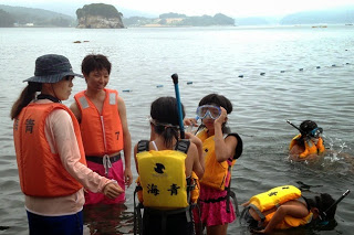 海辺の浅瀬で黄色やオレンジの救命胴衣を着た子どもたちがスノーケルの準備をしており、大人が見守る中で一人が水中に潜っている様子が見える写真