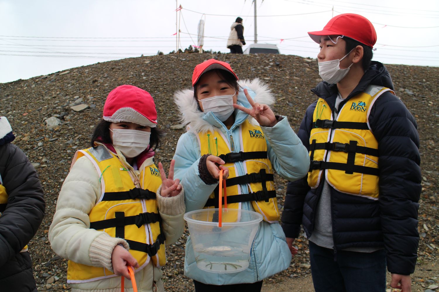 黄色のライフジャケットを身に着けそれぞれ稚魚が入ったバケツを手に持った生徒3人が、カメラにピースサインをしている写真