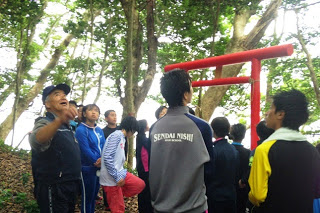 森の中の赤い鳥居の前に立ち、地元の案内人の話を聞く子どもたちの様子の写真