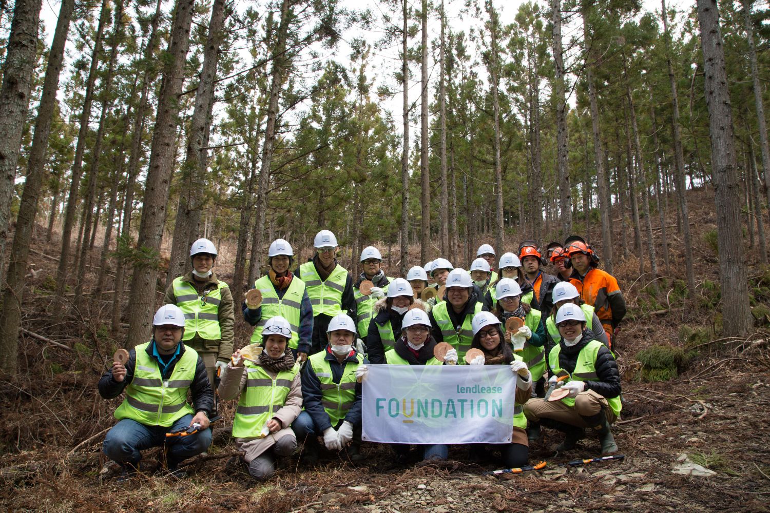 細い木々が生えた森の中で、蛍光ベストとヘルメットを着用したグループが、丸い木製オブジェを手に持ち並んでいる記念写真