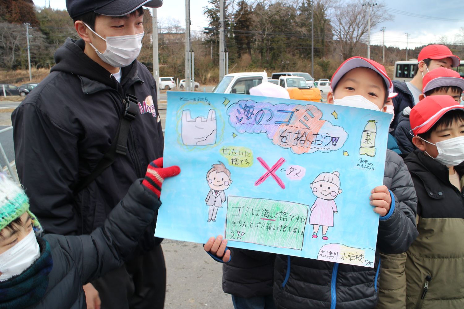 屋外で、マスクをつけた子どもたちが「海のゴミを拾おう」という標語とイラストが描かれた青色の手書きのポスターを持ってカメラに見せている写真