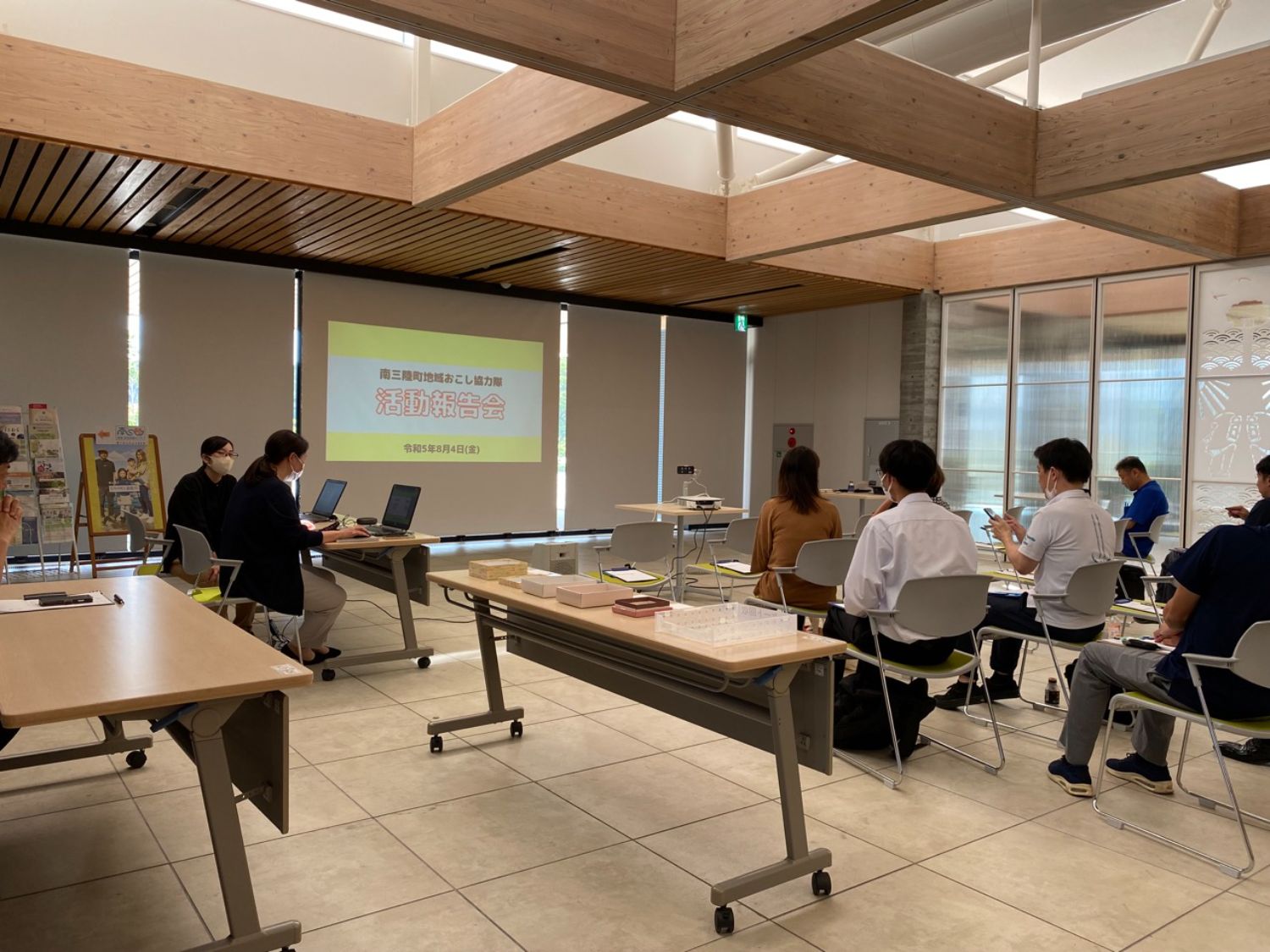 会議室で人々が椅子に座り、前方のスクリーンで「活動報告会」という文字が映し出され、活動報告会が行われている写真