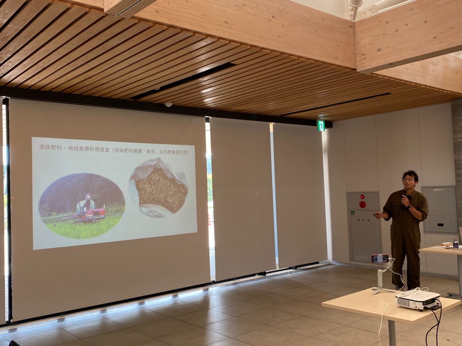木造の天井と白い壁のある会議室で、作業服姿の太田さんがマイクを持ってプロジェクターの前で発表をしている写真
