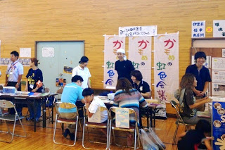 体育館内の壁際に設けられたブースで、「かもめの虹色談義」と書かれた垂れ幕の前に人が集まり、説明を受けたり話をしている写真