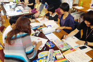 体育館内の机に並べられた色鉛筆やマーカーを使って、子どもと大人が一緒にイラストを描いている写真