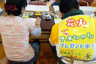 背中に「花火かアメちゃんプレゼント中！」と書かれた黄色い紙を貼った人が、隣の人と一緒に机に向かって絵を描いている写真