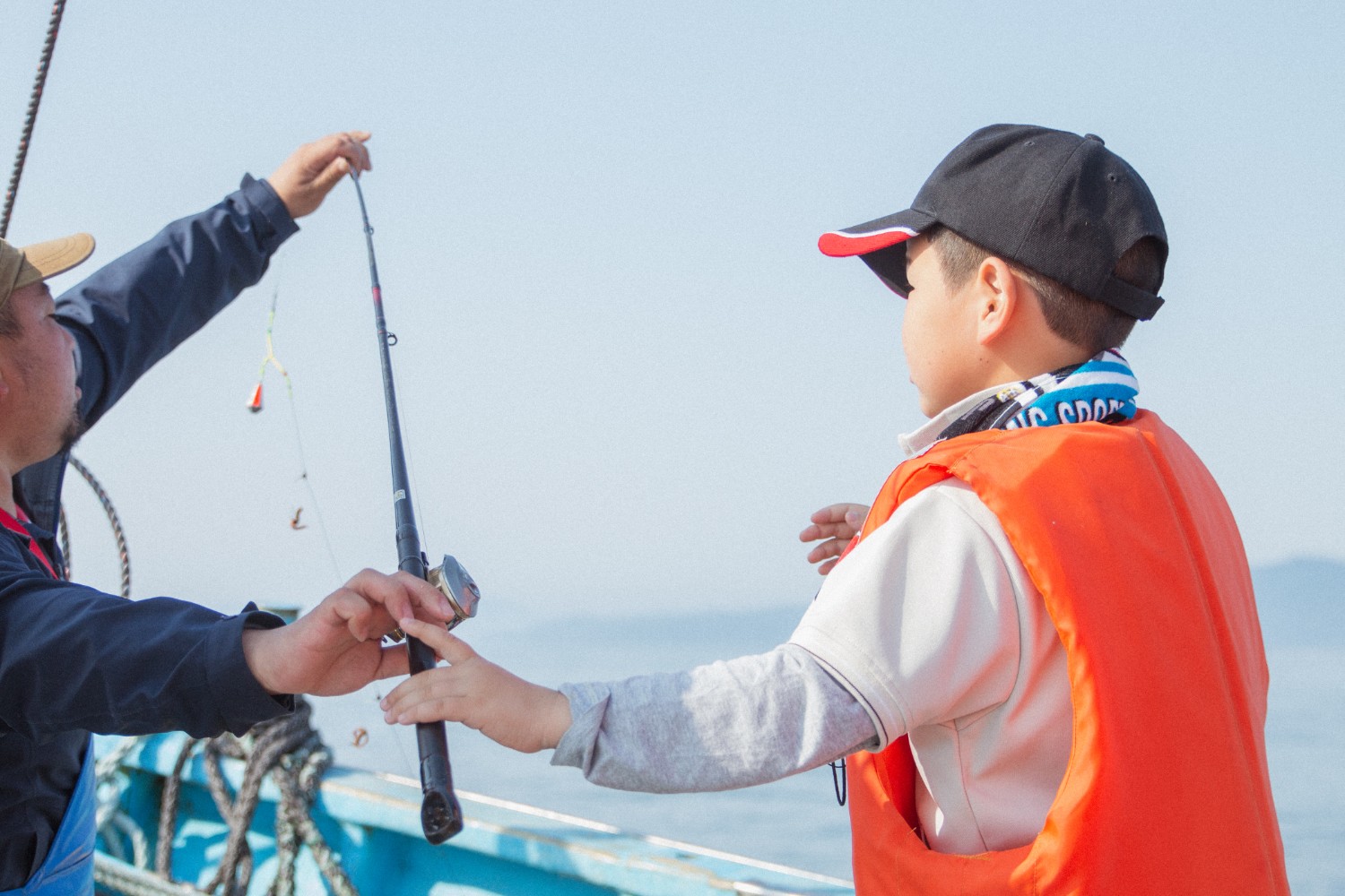 釣り竿を持っている青い作業服を着た父親からサポートを受けながら、黒い帽子とオレンジのライフジャケットを着た芳雅くんが釣り竿を手に取っている場面の写真