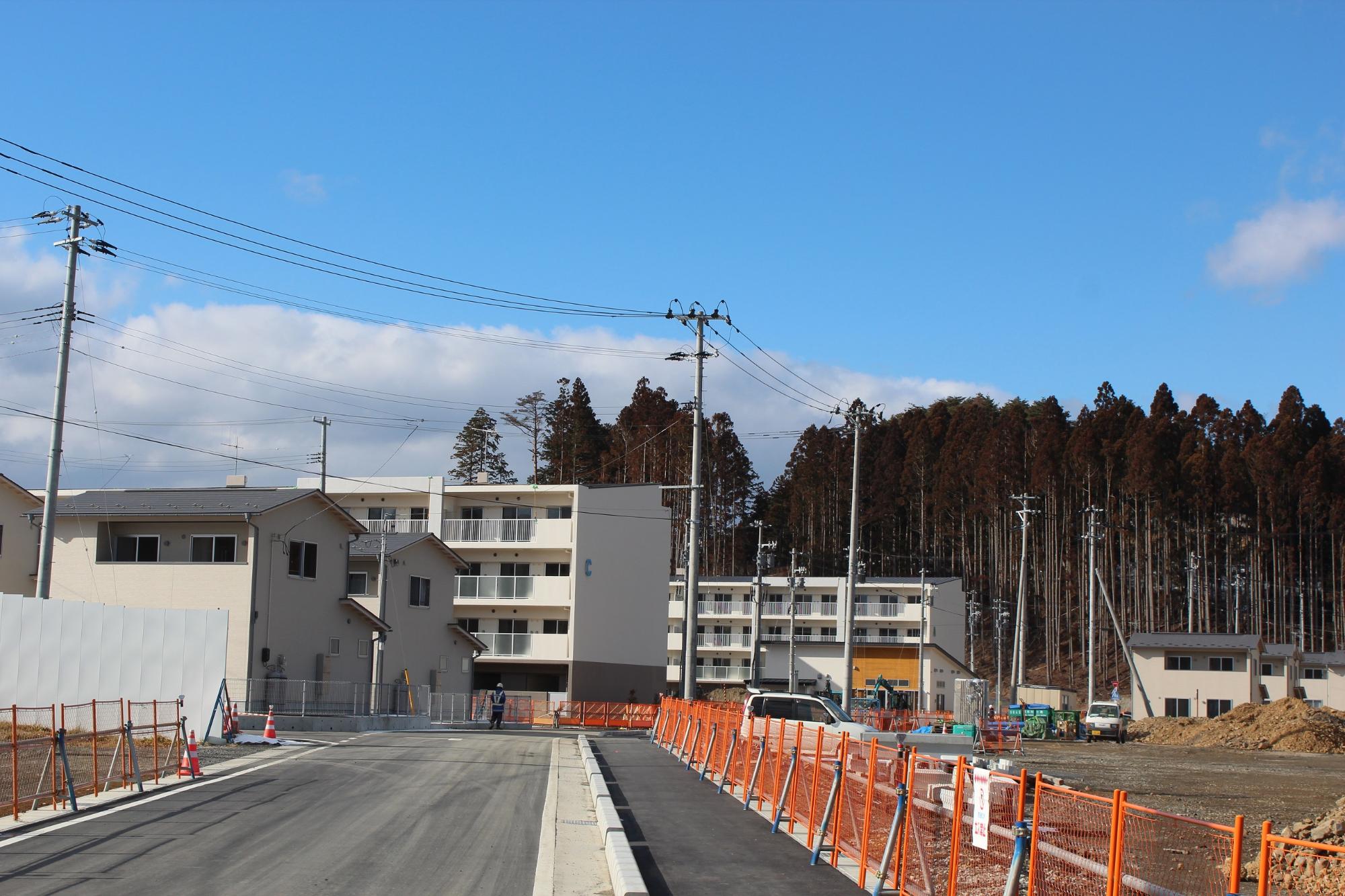 災害公営志津川中央住宅の建物がすべて外観を現しており、敷地内では舗装工事や植栽準備などの外構工事が進行中で、工事用車両や作業員が整備作業を行っている様子が映されている写真