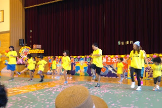 明るい黄色のTシャツを着た子どもたちと若者たちが床にピンク色の紙吹雪が多数散っている体育館のステージ前で音楽に合わせて楽しげに踊っている様子を写した写真