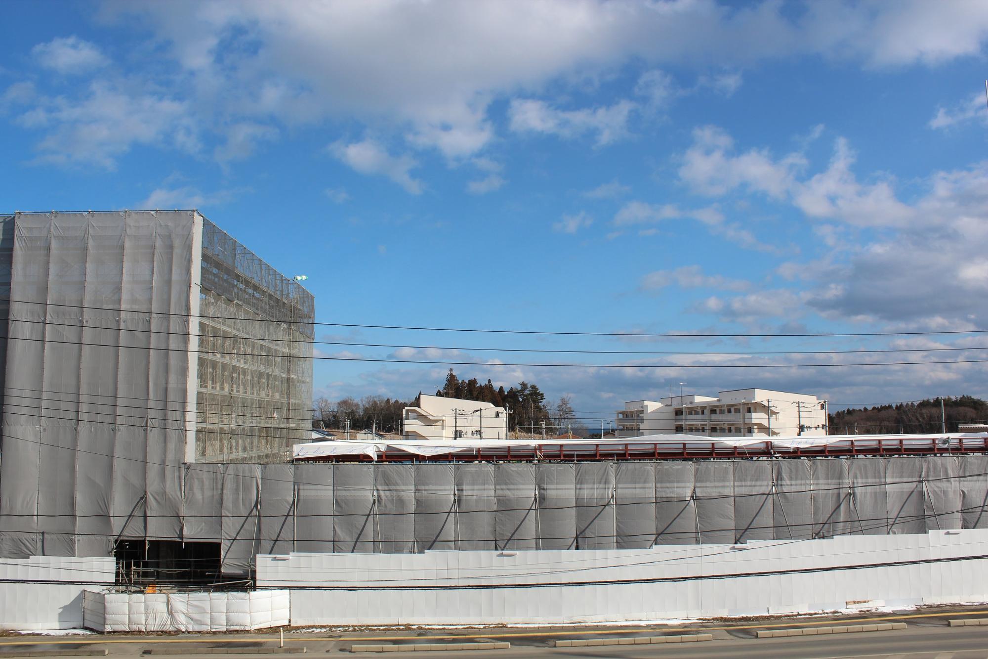 雲のかかった青空の下、建物の周囲には仮設のフェンスや工事用の資材が置かれている平成29年2月3日現在の写真
