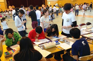 体育館内のイベントで、多くの人が集まる中、子どもたちが机で色ぬりやお絵かきをしている様子の写真